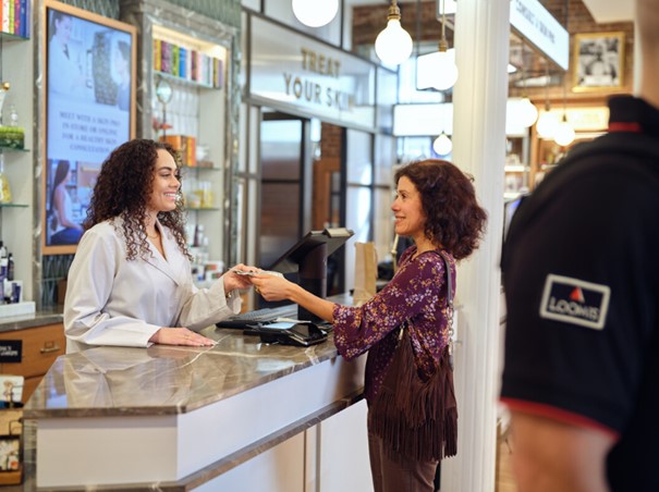 kvinne-betaler-i-kassa-i-en-butikk-til-smilende-ansatt-mens-loomisvekter-går-forbi-i-forgrunnen